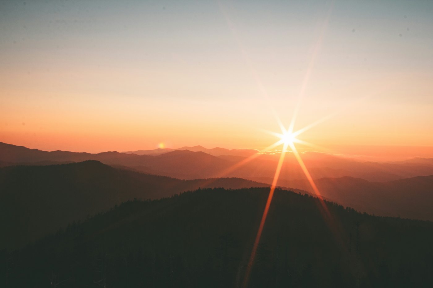 The sun setting over layers of misty mountain ridges, with rays of light across a clear orange sky